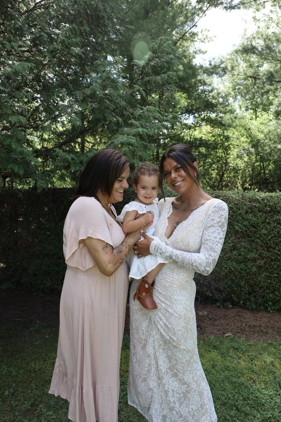A smiling mother in white lace holding her toddler, beside a woman in pink, in a leafy garden